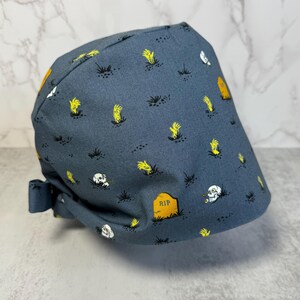May include: A dark blue surgical scrub cap with a repeating pattern of yellow zombie hands, tombstones, and skulls. The cap has a tie in the back for an adjustable fit. The tombstone has the letters "RIP" on it.