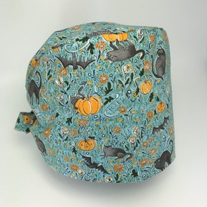 May include: A blue and green patterned scrub hat with black cats, pumpkins, and the words "Spooky Treats" printed on it.