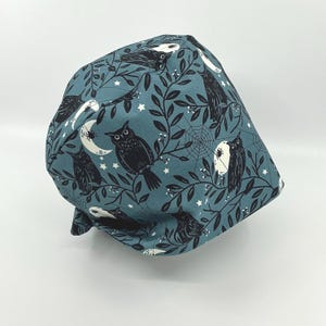 May include: A teal fabric cap featuring a repeating pattern of black owls, crescent moons, spiderwebs, and stars. The design is set against a backdrop of leafy vines. The cap is likely for medical professionals or those in the beauty industry.