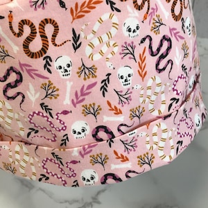 May include: A pink fabric item with a repeating pattern of orange and black snakes, white skulls, and bone shapes. The design also includes small sprigs of leaves and flowers in shades of purple and brown. The item has a folded brim.