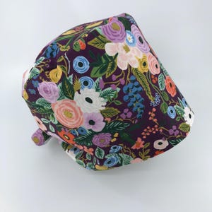 May include: Purple scrub hat with a floral print featuring pink, white, blue, and yellow flowers. The hat has a tie on the side.