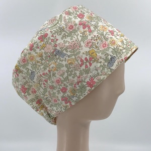 May include: A scrub cap featuring a cream-colored background with a repeating pattern of small, colorful flowers in shades of pink, yellow, and green, interspersed with tiny illustrations of cats in various poses and colors like grey, white, and orange.