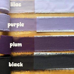 May include: A selection of fabric ribbons in various shades of purple and black, including lilac, purple, plum, and black. Each ribbon is labeled with its color name in white text. The ribbons have a frayed edge and are displayed on a wooden surface.