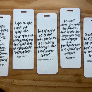 May include: Five white rectangular bookmarks with black handwritten script. Each bookmark features a different Bible verse, including Galatians 5:22, Psalm 130:7, 1 Corinthians 15:57, Psalm 91:4, and Isaiah 26:4. The bookmarks have a small hole at the top.