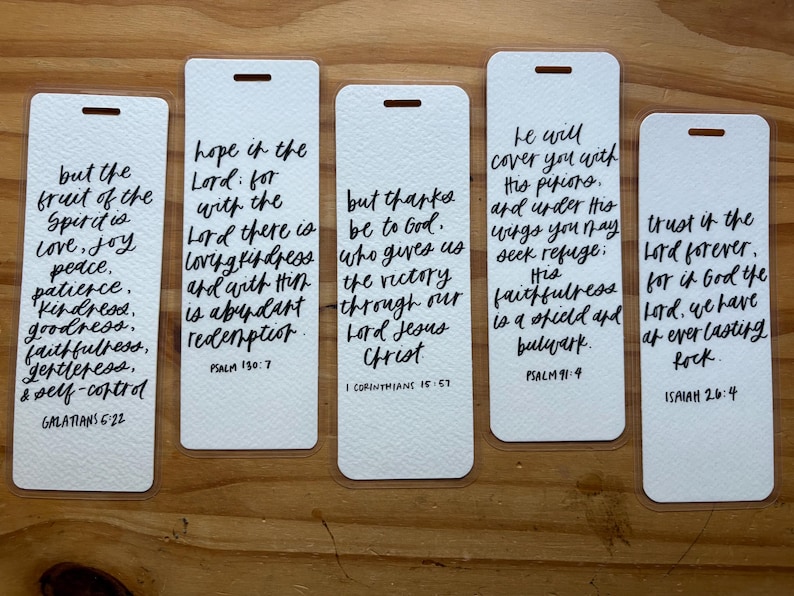 May include: Five white rectangular bookmarks with black handwritten script. Each bookmark features a different Bible verse, including Galatians 5:22, Psalm 130:7, 1 Corinthians 15:57, Psalm 91:4, and Isaiah 26:4. The bookmarks have a small hole at the top.