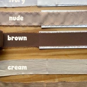 May include: A selection of fabric ribbons in various neutral colors, including ivory, nude, brown, cream, and sand. Each ribbon is labeled with its color name in white text. The ribbons have a frayed edge.