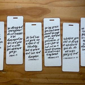 May include: Six white rectangular bookmarks with black handwritten script. Each bookmark features a different Bible verse. The bookmarks have a small hole at the top for a ribbon or string. The background is a wooden surface.