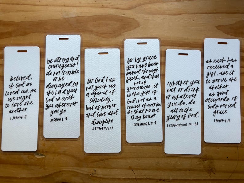 May include: Six white rectangular bookmarks with black handwritten script. Each bookmark features a different Bible verse. The bookmarks have a small hole at the top for a ribbon or string. The background is a wooden surface.