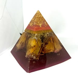 Może przedstawiać: Dekoracyjna piramida wykonana z przezroczystej żywicy, z głęboką fioletową podstawą i złotym wierzchołkiem. Suszone kwiaty są zawieszone w żywicy, dodając naturalny element do projektu. Kształt piramidy to klasyczna forma geometryczna.