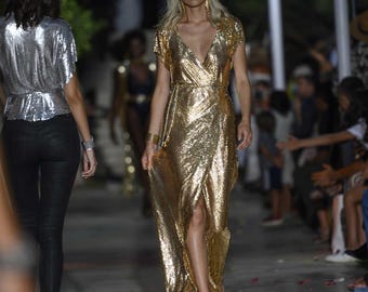 May include: A woman walks the runway wearing a gold sequined dress with a thigh-high slit. The dress has a V-neckline and short sleeves. She is wearing gold jewelry and has her blonde hair down.
