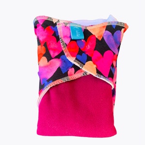 Può includere: Un assorbente riutilizzabile in tessuto foderato in pile rosa con sfondo nero e stampa ad acquerello di cuori colorati. L'assorbente ha un'etichetta blu con la scritta "Eudora".