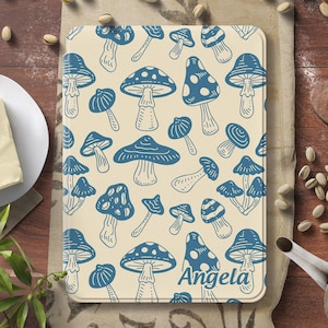 Könnte beinhalten: Ein rechteckiges Schneidebrett mit cremefarbenem Hintergrund und einem sich wiederholenden Muster aus blauen Pilzillustrationen. Der Name "Angela" ist unten in blauer Schrift aufgedruckt. Das Brett ist von einer Holzoberfläche umgeben.