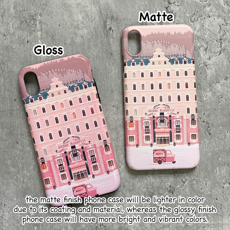 May include: Two phone cases, one glossy and one matte, featuring a pink building design with the text "GRAND BUDAPEST HOTEL". The matte case is lighter in color. A pink truck is visible on the bottom of the cases.