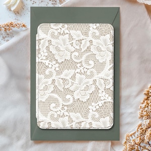 Può includere: Un motivo di pizzo bianco dettagliato su sfondo beige, inserito in una busta verde scuro. Il motivo floreale suggerisce un tema di eleganza, possibilmente per inviti o cancelleria.