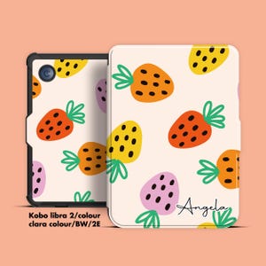 Könnte beinhalten: Eine Kobo Libra 2 E-Reader-Hülle mit cremefarbenem Hintergrund und einem sich wiederholenden Muster aus bunten Erdbeeren. Die Erdbeeren sind orange, gelb und rosa mit grünen Blättern. Der Name "Angela" ist handschriftlich auf der Hülle.