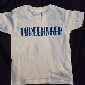 May include: White t-shirt with the text "Three-nager" in blue and green lettering.