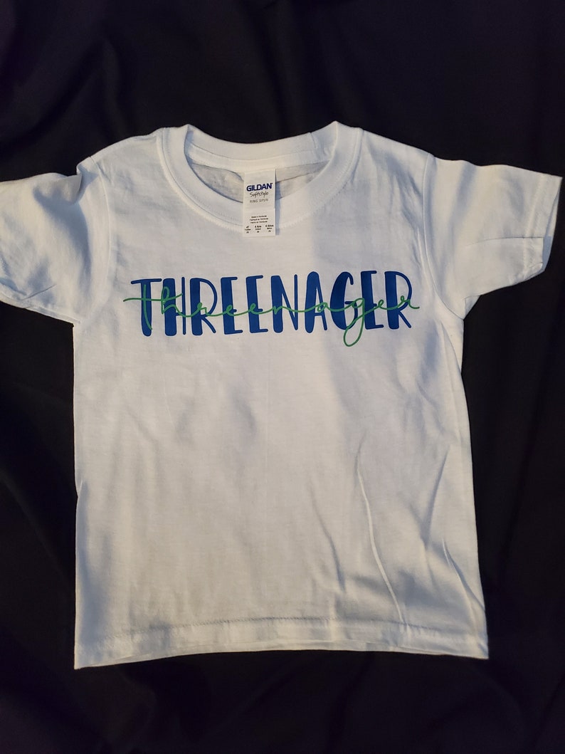 May include: White t-shirt with the text "Three-nager" in blue and green lettering.