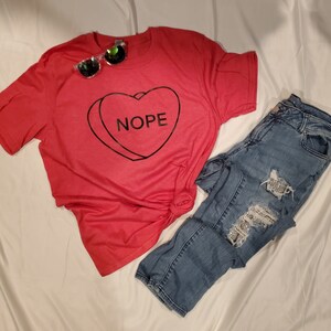 Puede incluir: Camiseta roja con un diseño de corazón negro que dice "NOPE". La camiseta se combina con unos vaqueros azules.