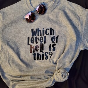 Puede incluir: Camiseta gris con texto negro que dice "¿Qué nivel de infierno es este?" Un par de gafas de sol rosas están encima de la camiseta.