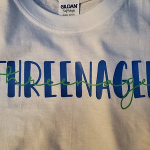 May include: White t-shirt with the text "THREEnager" in blue and green lettering.