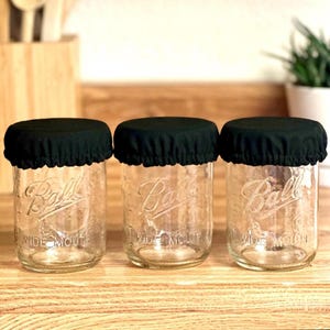 May include: Three clear glass Ball Wide Mouth jars with black fabric covers. The jars are sitting on a wooden surface. The jars are empty and ready to be filled. The background includes a wooden cutting board and a plant in a white pot.