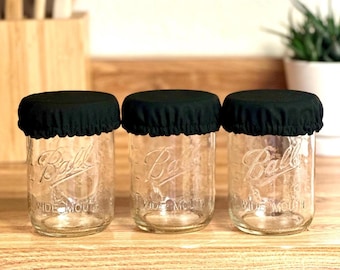 May include: Three clear glass Ball Wide Mouth jars with black fabric covers. The jars are sitting on a wooden surface. The jars are empty and ready to be filled. The background includes a wooden cutting board and a plant in a white pot.