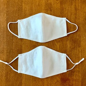 Two white face masks with elastic ear loops. The masks are made of a soft, breathable fabric and have a simple design.