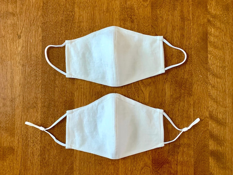Two white face masks with elastic ear loops. The masks are made of a soft, breathable fabric and have a simple design.