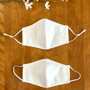 May include: Two white cotton face masks with elastic ear loops. The masks are plain and have a simple design.