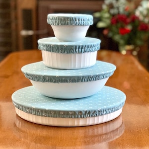 May include: A set of four blue and white reusable bowl covers with a floral pattern. The covers are made of fabric and have a ruffled edge. They are stacked on top of each other on a wooden table.