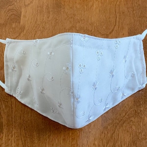 May include: White face mask with white embroidered floral pattern. The mask has elastic ear loops.