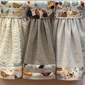May include: Three hanging kitchen towels with decorative chicken-themed fabric accents. The towels have different textures and colors, including cream, tan, and white. The fabric borders feature various chicken and farm-themed patterns, adding a rustic touch to the kitchen decor.