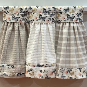 May include: Three hanging kitchen towels with a butterfly theme. Each towel has a different texture: waffle, ribbed, and striped. The top and bottom borders feature a butterfly and floral pattern in black, white, and pink.