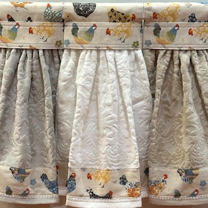 May include: Three hanging kitchen towels featuring a cream-colored fabric band with a colorful chicken and floral print at the top and bottom. The center sections of the towels are textured with a raised damask pattern in beige and white.