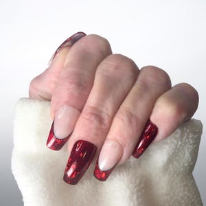 Puede incluir: Mano con uñas artificiales largas y cuadradas. Las uñas presentan un diseño de manicura francesa con base transparente y punta roja brillante. La mano sostiene una toalla blanca y esponjosa.