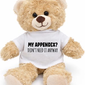 May include: A plush teddy bear with light brown fur, black eyes, and a brown nose. The bear wears a white t-shirt with the text "MY APPENDIX? DIDN'T NEED IT ANYWAY" in black. The bear is sitting upright against a white background.