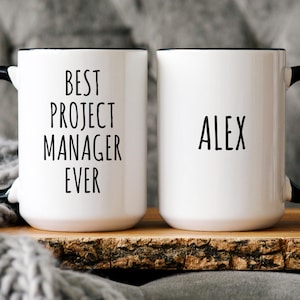May include: Two white ceramic mugs with black handles and rims. One mug reads "BEST PROJECT MANAGER EVER" in black text. The other mug has the name "ALEX" printed in black. The mugs are on a wooden tray.
