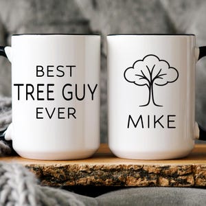 May include: Two white ceramic mugs with black handles and rims. One mug reads "BEST TREE GUY EVER" in black text. The other mug has a tree graphic and the name "MIKE". Both mugs are on a wooden tray.