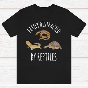 May include: Black t-shirt with white text that reads "Easily Distracted By Reptiles". The shirt features a cartoon image of a snake, a lizard, and a turtle.