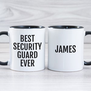 May include: Two white ceramic mugs with black handles and black rims. One mug says "Best Security Guard Ever" and the other mug says "James".