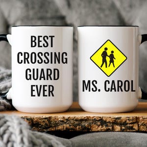 May include: White ceramic mug with a black handle and rim. The mug features the text "BEST CROSSING GUARD EVER" on one side and a yellow pedestrian crossing sign with the name "MS. CAROL" on the other.