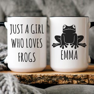 May include: Two white ceramic mugs with black handles and rims. One mug reads "JUST A GIRL WHO LOVES FROGS" in black text. The other mug features a black frog illustration and the name "EMMA".