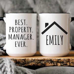 May include: Two white ceramic mugs with black handles and rims. One mug reads "BEST. PROPERTY. MANAGER. EVER." in black text. The other mug features a black house graphic and the name "EMILY" in black text. Both mugs are on a wooden tray.