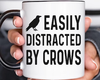 May include: White ceramic mug with a black rim and handle. The mug has a black silhouette of a crow and the text "EASILY DISTRACTED BY CROWS".