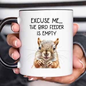 May include: White ceramic mug with a black handle. The mug has a squirrel illustration and the text "Excuse me... the bird feeder is empty".