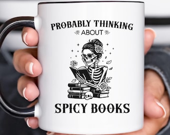May include: White ceramic mug with a black interior and handle. The mug features a black and white illustration of a skeleton reading a book, with the text "PROBABLY THINKING ABOUT SPICY BOOKS".