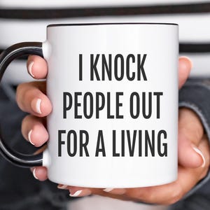 May include: White ceramic mug with black interior and handle. The mug features the text "I KNOCK PEOPLE OUT FOR A LIVING" in bold black letters. The mug is held by a person.