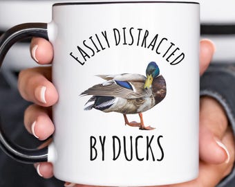 May include: White ceramic mug with a black handle and black rim. The mug has the text "Easily Distracted By Ducks" printed in black. A cartoon image of a duck is printed in the center of the mug.