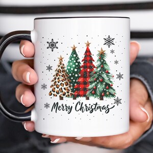 May include: White ceramic mug with a black handle. The mug features a Christmas design with three stylized Christmas trees in red, green, and leopard print. The trees are decorated with ornaments and snowflakes. The text "Merry Christmas" is printed below the trees.