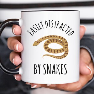 May include: White ceramic mug with a black handle and rim, featuring the text "EASILY DISTRACTED BY SNAKES" and a brown snake illustration. The mug is held by a person.